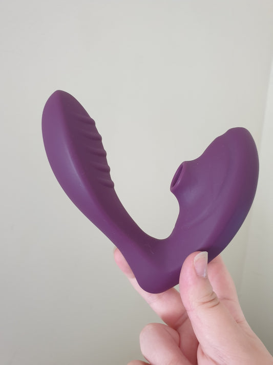Purple suction vibrator held in one hand against a plain light wall, showing the ribbed curved arm and the round suction opening.