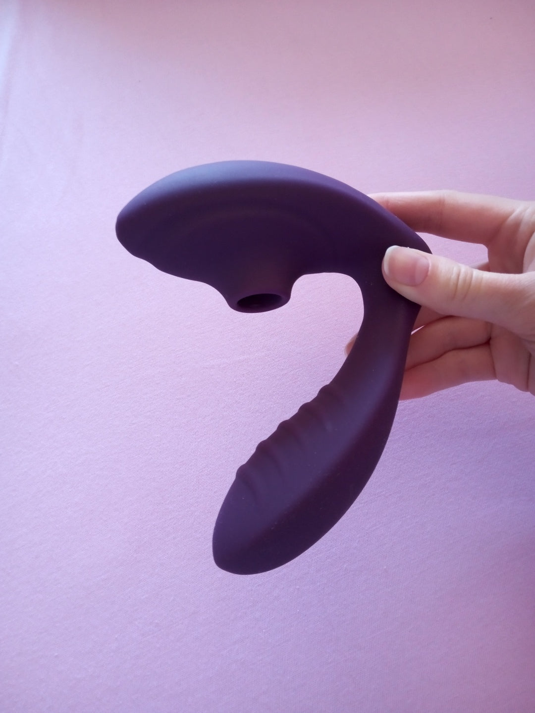 Close-up of a purple suction vibrator held up against a lilac backdrop, showing the round suction opening on the head and the ribbed curved arm.